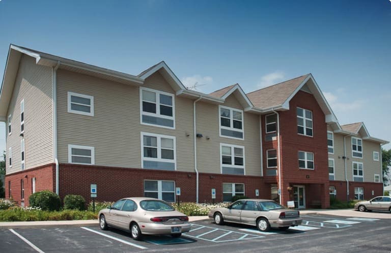 A two-story building with beige siding and red brick accents is shown. Several cars are parked in the lot in front of the building, exemplifying affordable housing in Bloomington, Indiana.