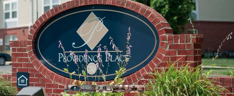 A sign for "Providence Place," representing affordable housing in Bloomington, Indiana, with a decorative logo, mounted on a red brick structure, surrounded by decorative grasses and plants.