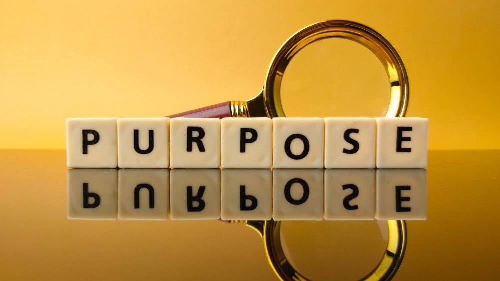 The word "PURPOSE" is spelled out in block letters reflected on a yellow surface with a magnifying glass in the background.