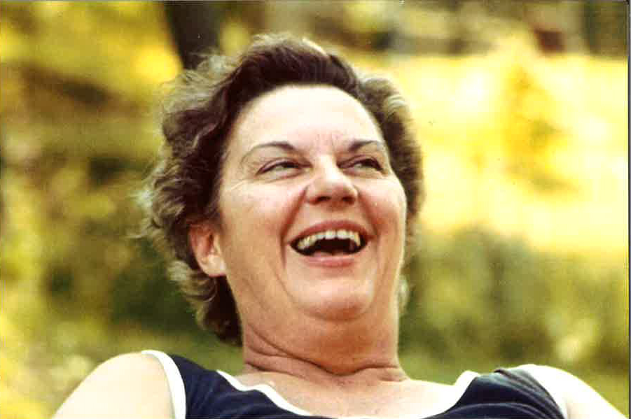 A middle-aged woman with short hair wearing a sleeveless top is smiling and laughing outdoors on a sunny day.