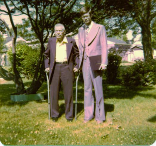 Two men standing outdoors on a grassy lawn; one older man with crutches in a suit, and one taller man holding a book or folder, also in a suit. Trees and houses are visible in the background.