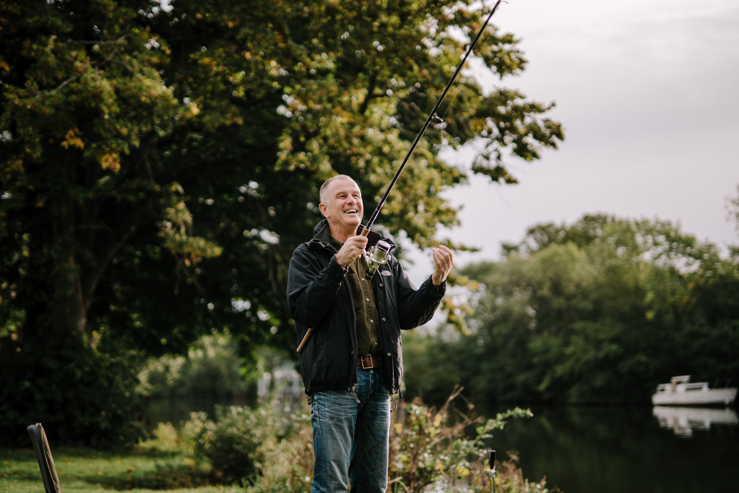 A person stands near a river, holding a fishing rod and appearing to have caught a fish. Dense trees and greenery form a serene backdrop, evoking the peaceful simplicity that comes with rightsizing for older adults.