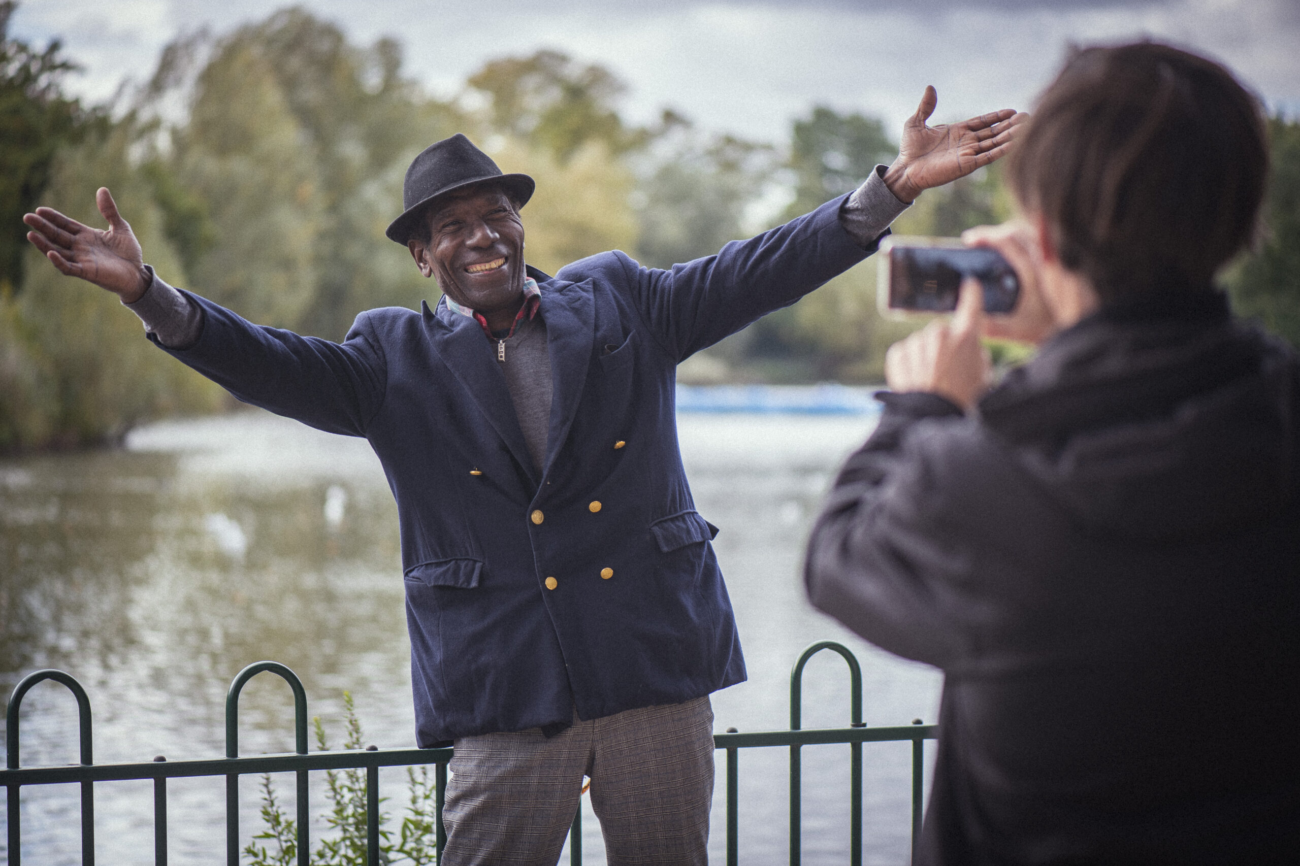 A man in a suit and hat smiles with arms wide open near a lake while another person takes his photo using a smartphone. Trees and water are visible in the background.