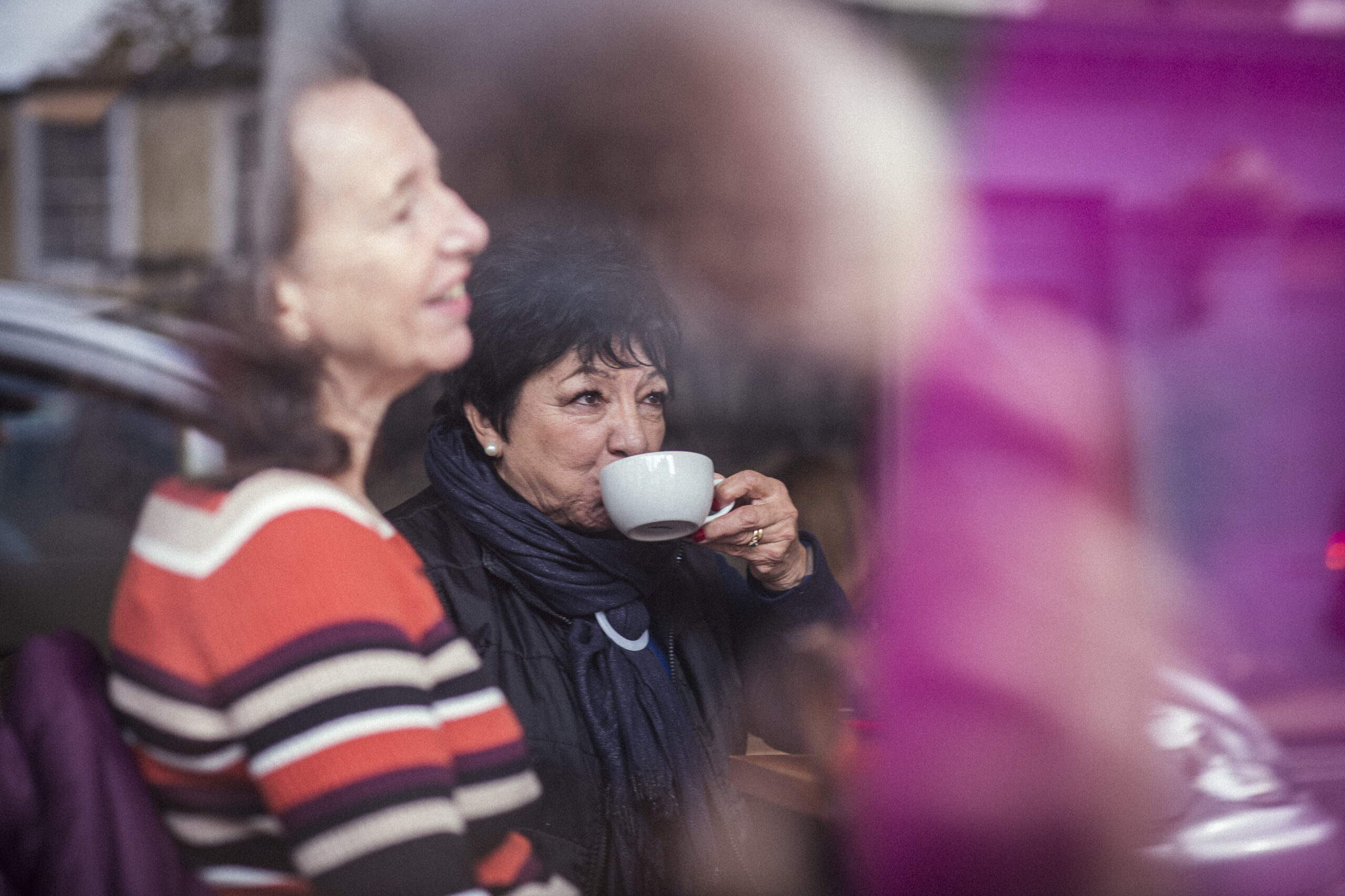 Two women are sitting in a cafe, one sipping from a cup while the other smiles. The image is partially obscured by reflections on the window.