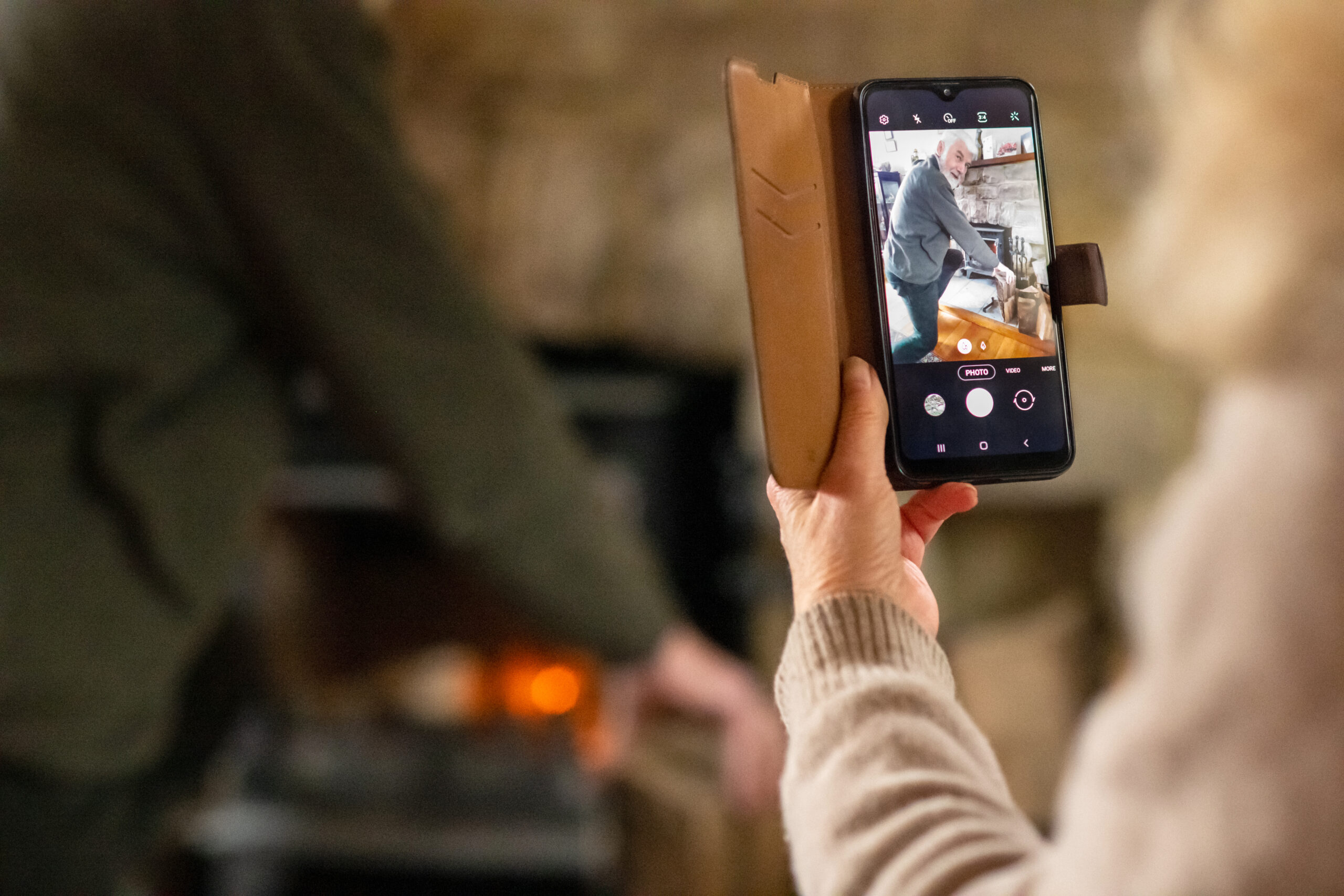 Person holding a smartphone, capturing another person putting wood into a fireplace.