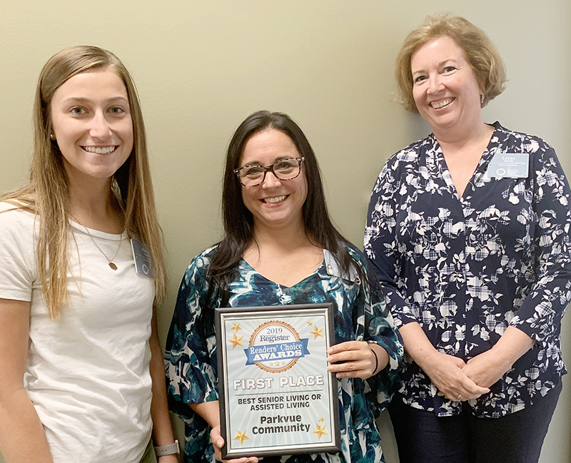 Three women stand smiling against a wall. The woman in the center holds a plaque that reads “First Place Best Senior Living or Assisted Living Parkvue Community.”.