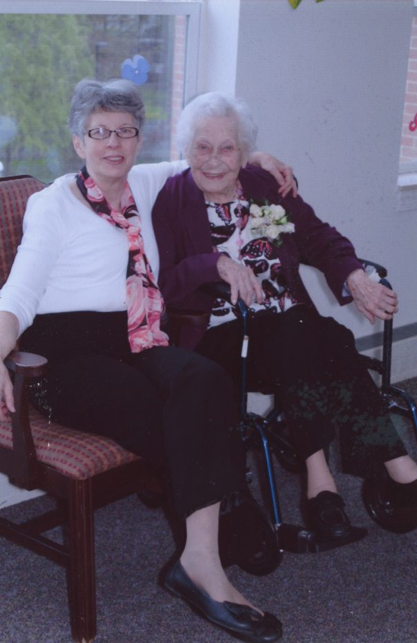 Two elderly women sit together, one on a chair and the other in a wheelchair, both smiling. The woman on the left has her arm around the woman on the right.