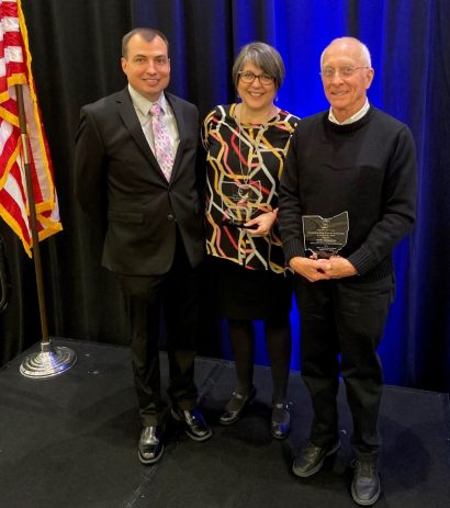 Three people stand in front of a black curtain, with an American flag in the background. Two of them hold awards, posing for a photo.