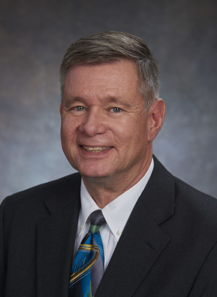 A middle-aged man with short gray hair, smiling confidently, wearing a dark suit, white shirt, and a multicolored tie, poses for a professional headshot that exudes leadership.