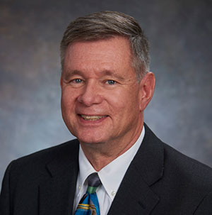 A man with short grey hair, wearing a dark suit and tie, smiles in front of a neutral background, following his announcement as the appointed President in the leadership restructure.