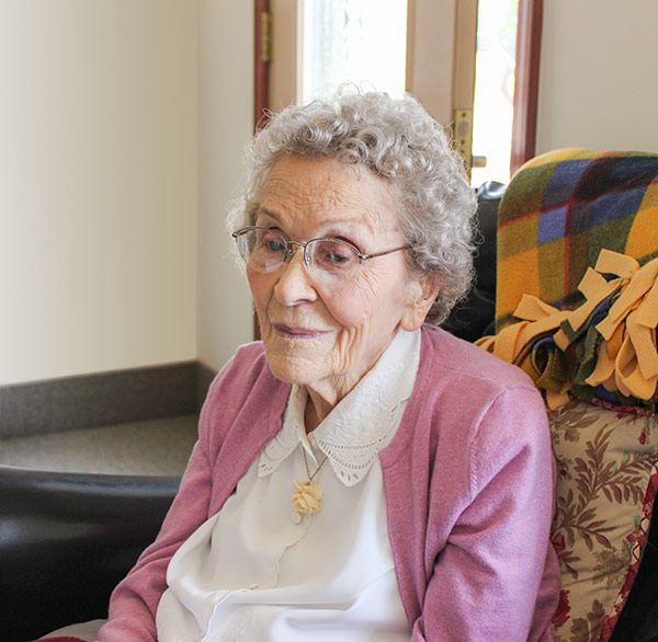 An elderly woman with curly gray hair, glasses, and a pink cardigan sits in a chair with a colorful blanket draped over it. She is in a well-lit room by a window.