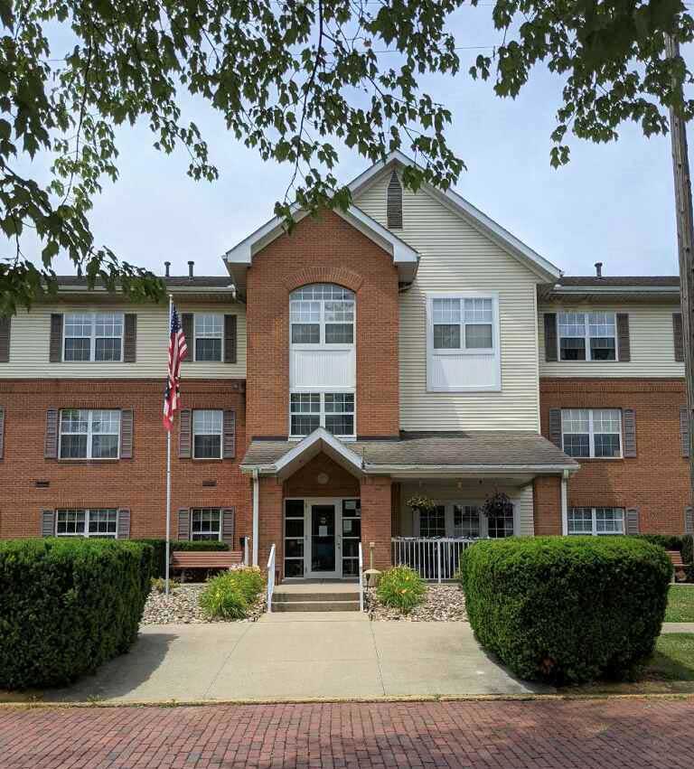 A three-story brick and siding residential building offering affordable housing in Brilliant, Ohio, with an American flag outside, flanked by manicured bushes and trees. The entrance features a small ramp and steps leading up to double glass doors.