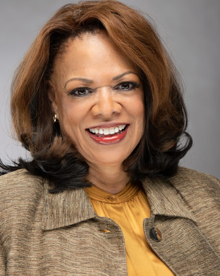 A smiling woman with shoulder-length brown hair, embodying leadership, wears a beige blazer over a yellow blouse as she poses against a gray background.