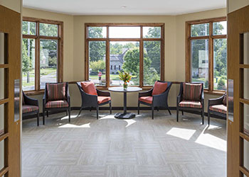 A sunlit room with large windows, five chairs arranged around a table with a flower vase, and parquet flooring.