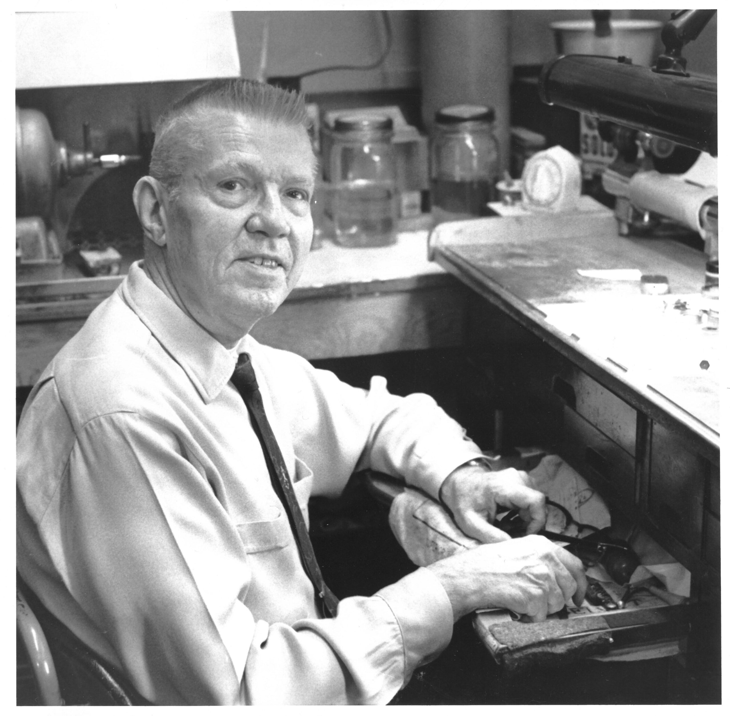 A man in a shirt and tie sits at a workbench, handling tools in a workshop with various equipment and jars in the background.