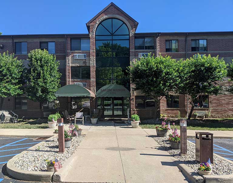 A brick residential building with a peaked glass entrance surrounded by trees and flower beds, viewed from a paved walkway. This charming example of affordable housing in Fort Wayne, Indiana offers both beauty and accessibility.