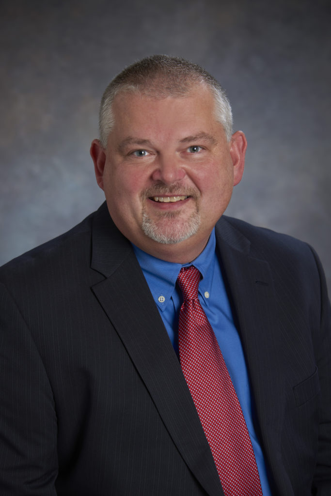 A man with a short beard and grey hair in a suit, blue shirt, and red tie, exuding leadership as he smiles against a gray background.