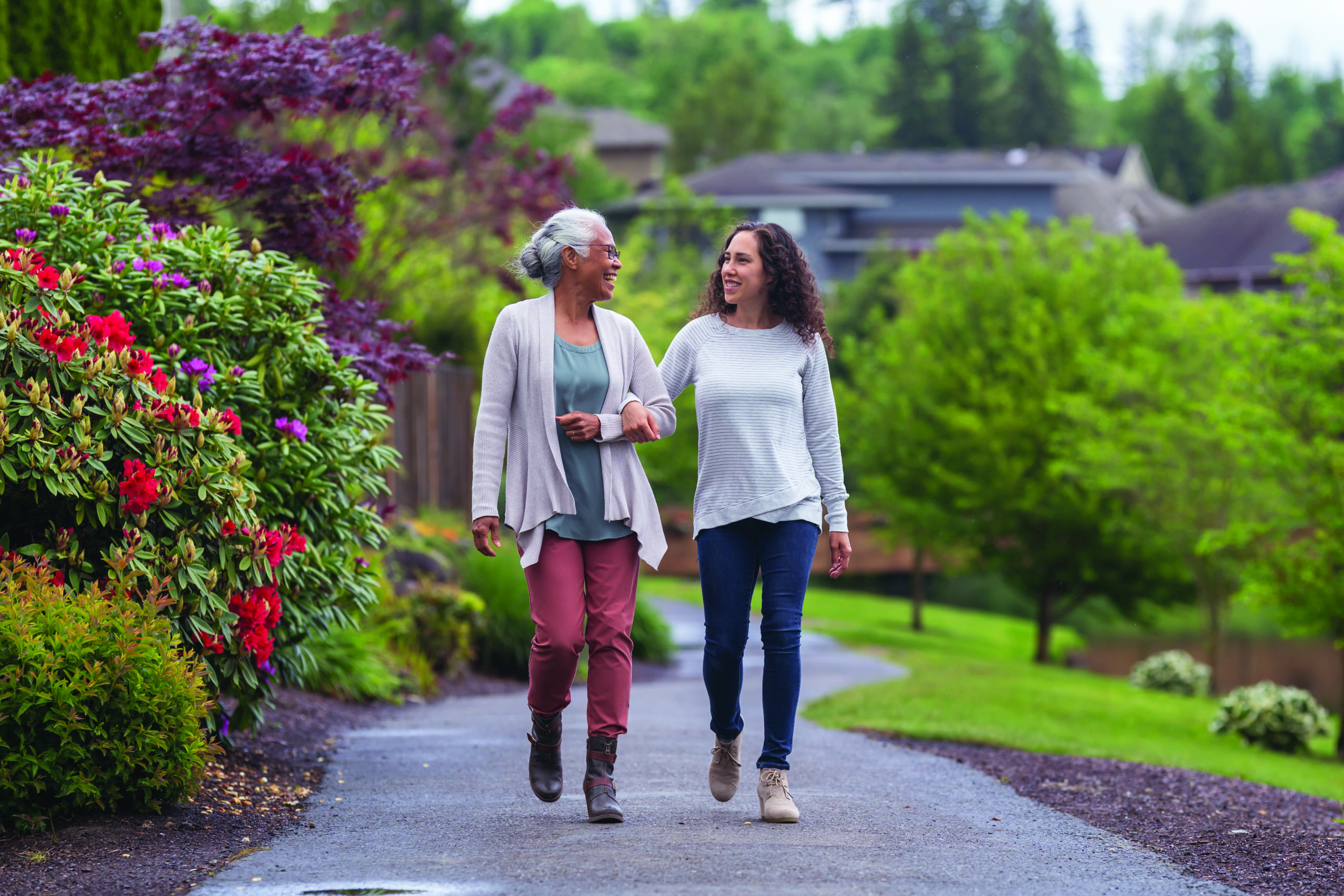 Two women walk arm-in-arm on a garden path, surrounded by lush greenery and vibrant flowers, with houses visible in the background. This scene embodies how service coordination extends care beyond communities, nurturing both environment and relationships.