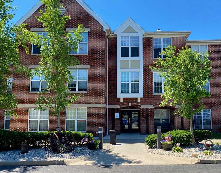 A three-story red brick building with multiple windows, a central entrance, and a gabled roof stands as a testament to affordable housing in Bellefontaine, Ohio. The front area features shrubs, decorative items including small flags, and two trees under a clear blue sky.