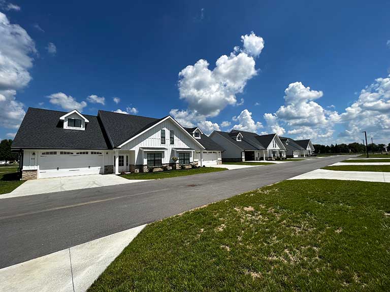 A neighborhood with several modern single-story homes lined up along a wide residential street under a partly cloudy sky. Each house has a driveway and a small front lawn.