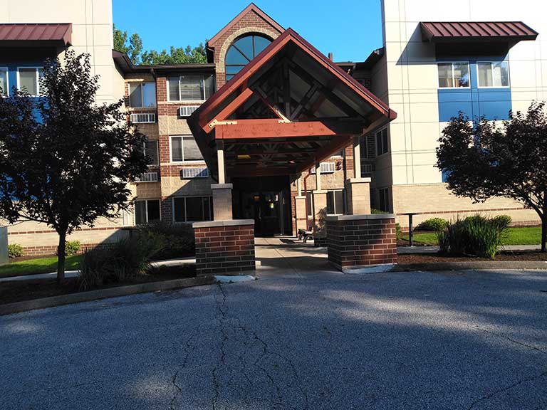 The front entrance of a multi-story building with a triangular glass canopy is surrounded by trees and a paved driveway, offering inviting access to affordable housing in Bedford, Ohio.