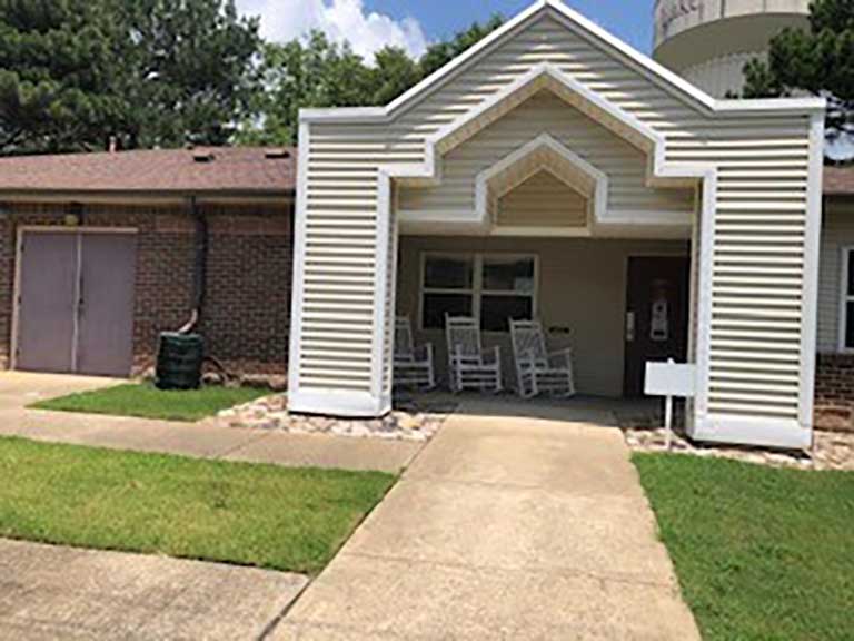 A building with a covered entrance featuring two rocking chairs, adjacent to a grassy lawn and a water tower in the background, exemplifies affordable housing in Horn Lake, Mississippi.