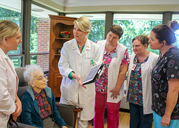 A group of healthcare professionals in white coats and scrubs gather around an elderly woman seated in a chair, discussing documents. The setting appears to be a well-lit room with large windows.