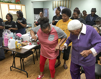 A group of people engage in a lively dance in a community room, with tables and various items in the background.