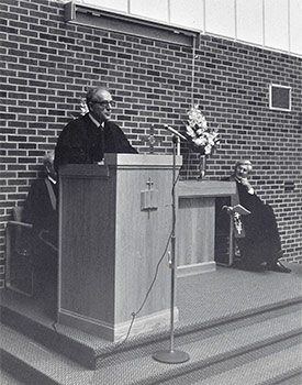 A man stands at a podium speaking into a microphone in what appears to be a church setting. Two other individuals are seated nearby.