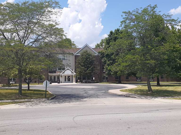 A brick building with a covered entrance, part of the affordable housing in Delaware, Ohio, is surrounded by trees and greenery. It features a curved driveway with a street in the foreground under a partly cloudy sky.