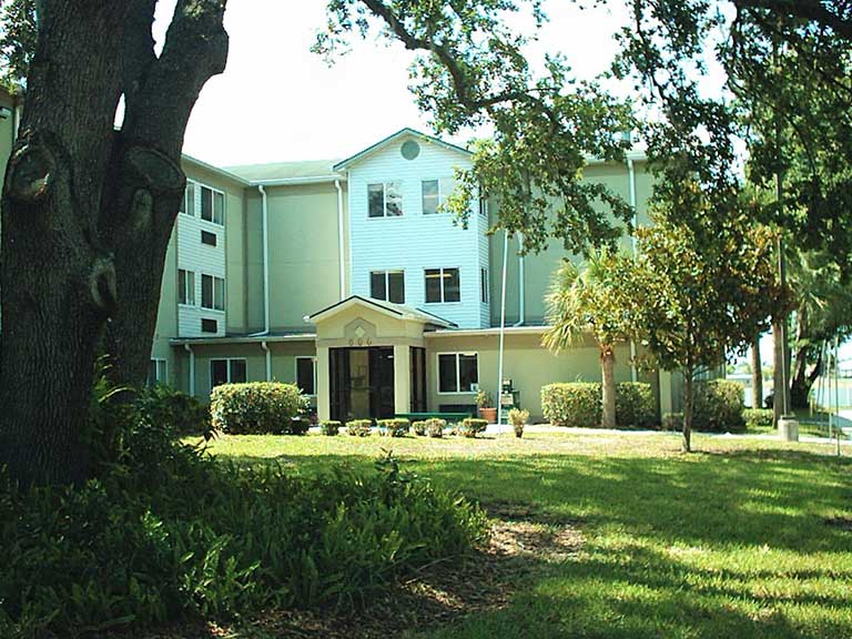 A three-story light green residential building with a covered entrance is surrounded by trees and manicured shrubs. The lawn in the foreground is shaded by large trees, offering serene affordable housing in Lakeland, Florida.