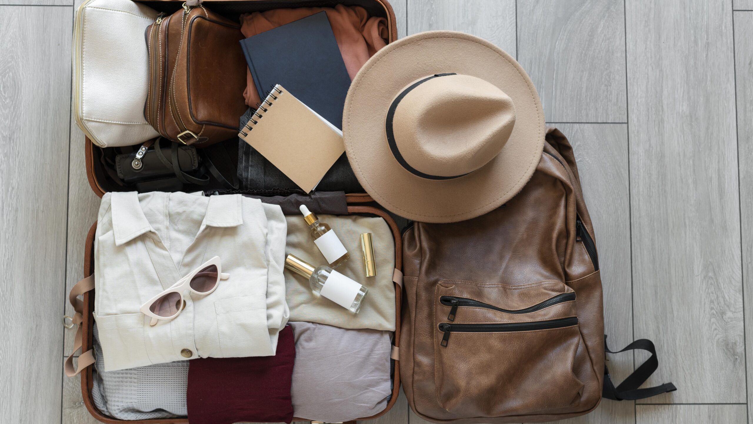 Open suitcase with clothing, sunglasses, a notebook, and toiletries next to a brown backpack and a tan hat on a wooden floor.