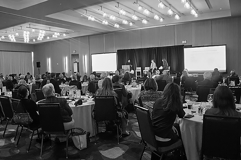 A black and white photo of attendees seated at round tables, listening to a panel of three speakers discussing technology and aging on a stage in a conference room with a projected screen in the background.