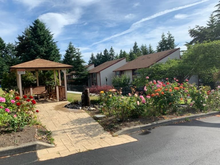 A landscaped garden with colorful flowers, a fountain, and a wooden gazebo with benches next to affordable housing in Tacoma, Washington, under a partly cloudy sky.