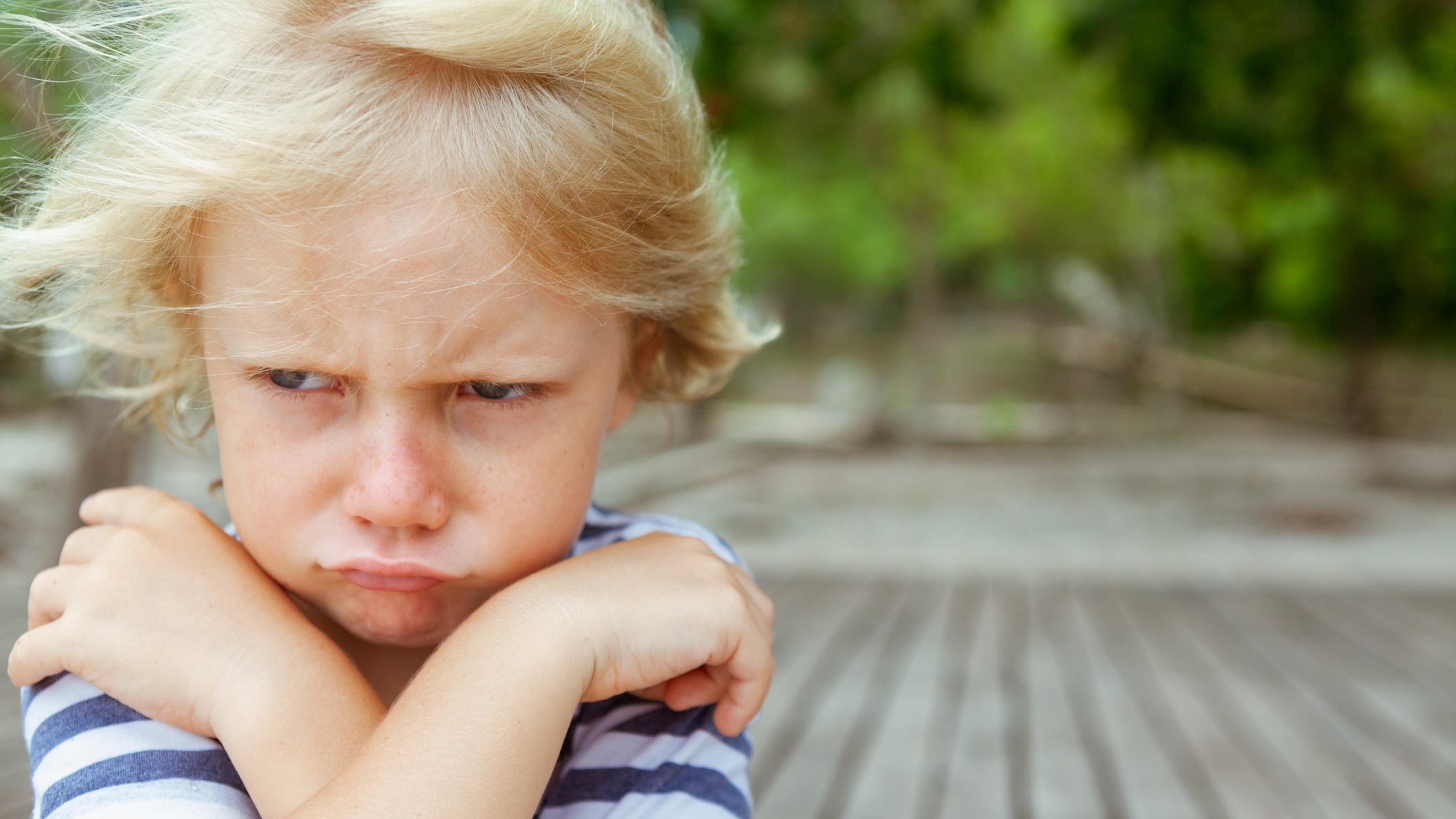 A young child with short blond hair and a striped shirt appears upset, crossing their arms and pouting. The background is a blurred outdoor setting with wooden planks and greenery.