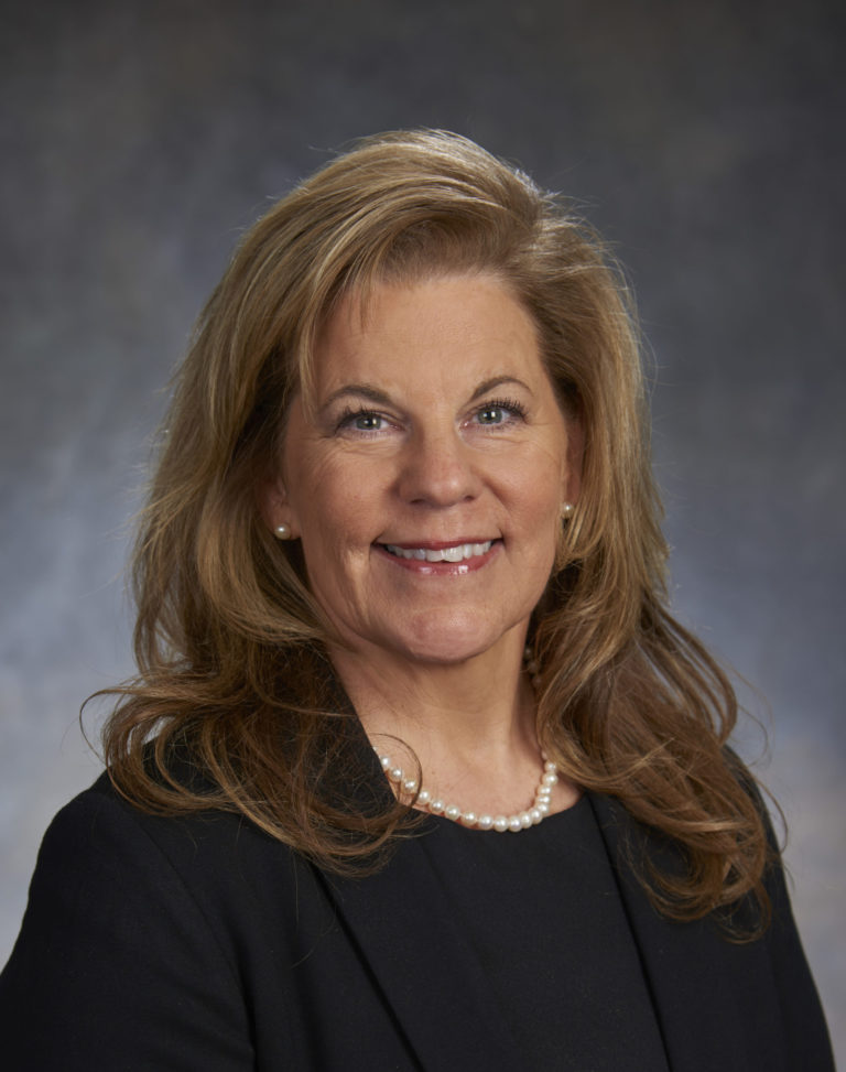 A woman with long blonde hair, wearing a black blazer and pearl necklace, exudes leadership as she smiles confidently against a dark, blurred background.