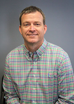 A man in a checkered shirt sits against a plain blue background. He has short hair and is smiling subtly.