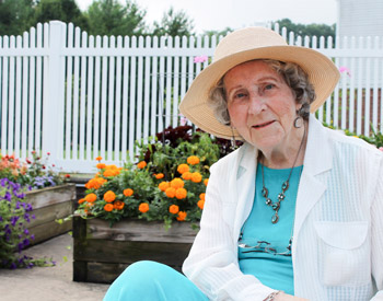 Elderly woman in a wide-brimmed hat and white outfit sits in front of a white picket fence with colorful flowers in planter boxes.