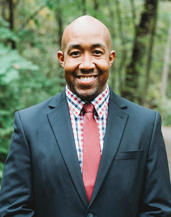 A man wearing a dark suit, checkered shirt, and red tie smiles while standing outdoors with trees in the background.