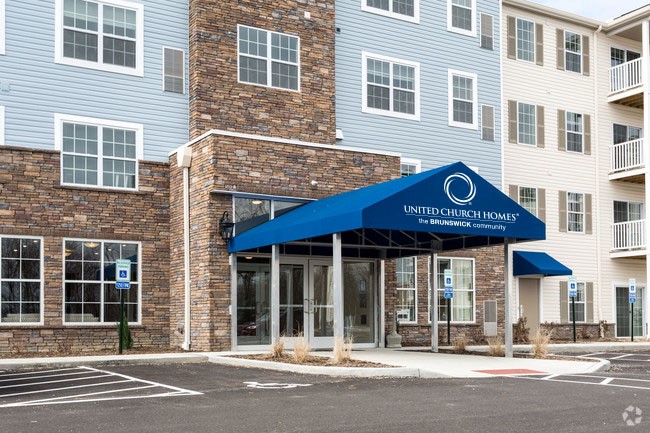 Entrance of a multi-story building with a blue canopy that reads "United Church Homes: The Brunswick Community" and accessible parking spaces in front.