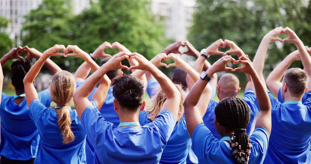 A group of people in blue shirts stands outdoors with their backs to the camera, forming heart shapes with their hands held above their heads as part of a campaign for abundant life.