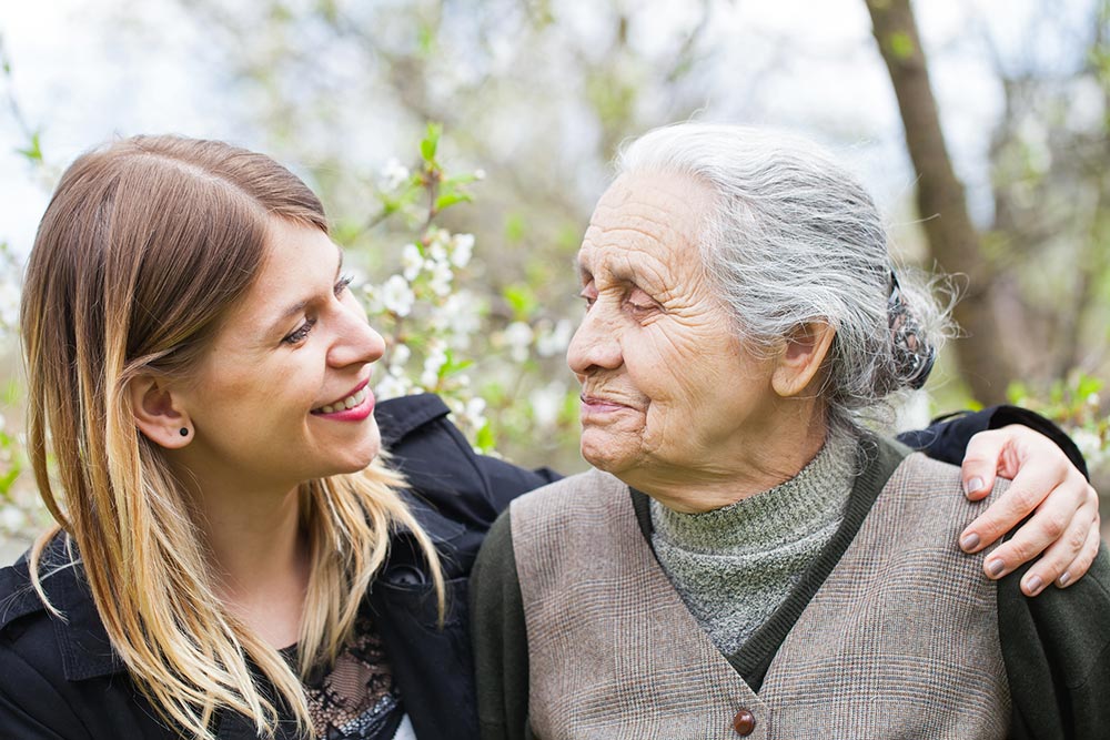 A young woman and an elderly woman share a moment together, with the younger woman smiling and the older woman looking in her direction in an outdoor setting, reflecting the importance of quality memory care services for fostering meaningful connections.