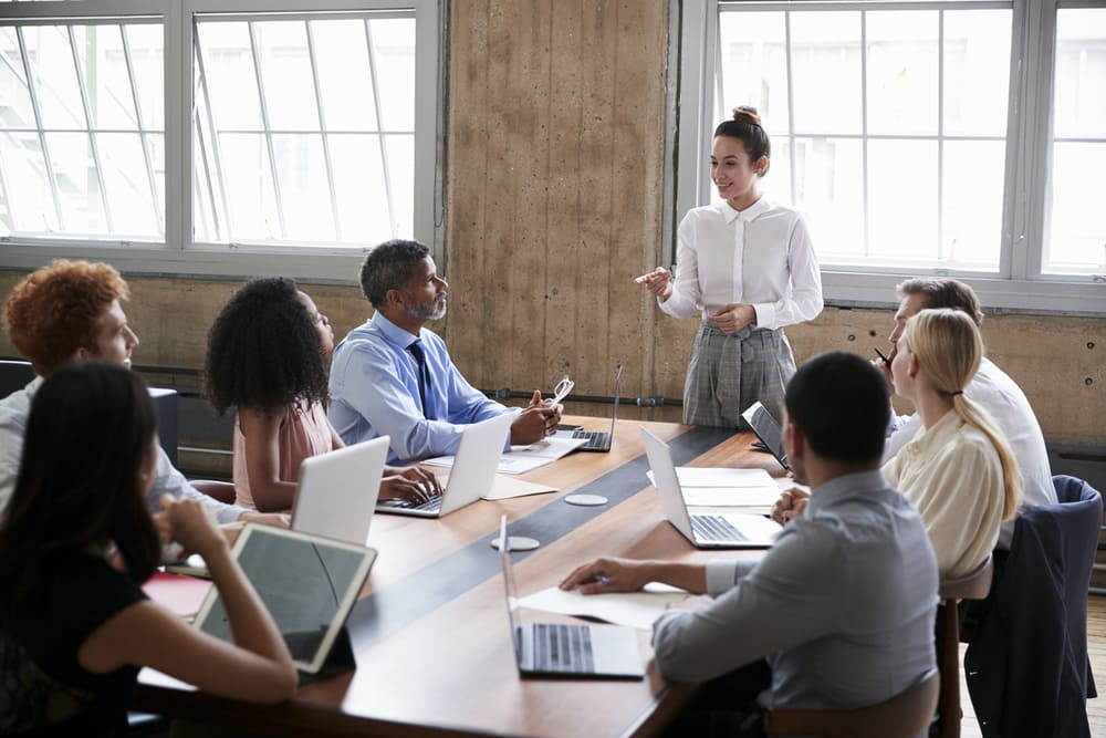 A woman in a white shirt stands and speaks to a group of seven seated colleagues around a conference table with laptops and documents, discussing careers at United Church Homes. They are in a bright room with large windows and concrete walls.