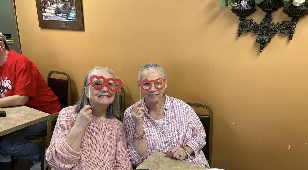 Two elderly women sitting at a table, smiling, and holding heart-shaped eyeglasses to their faces indoors with beige walls and a painting in the background, enjoying their time in affordable housing near you.