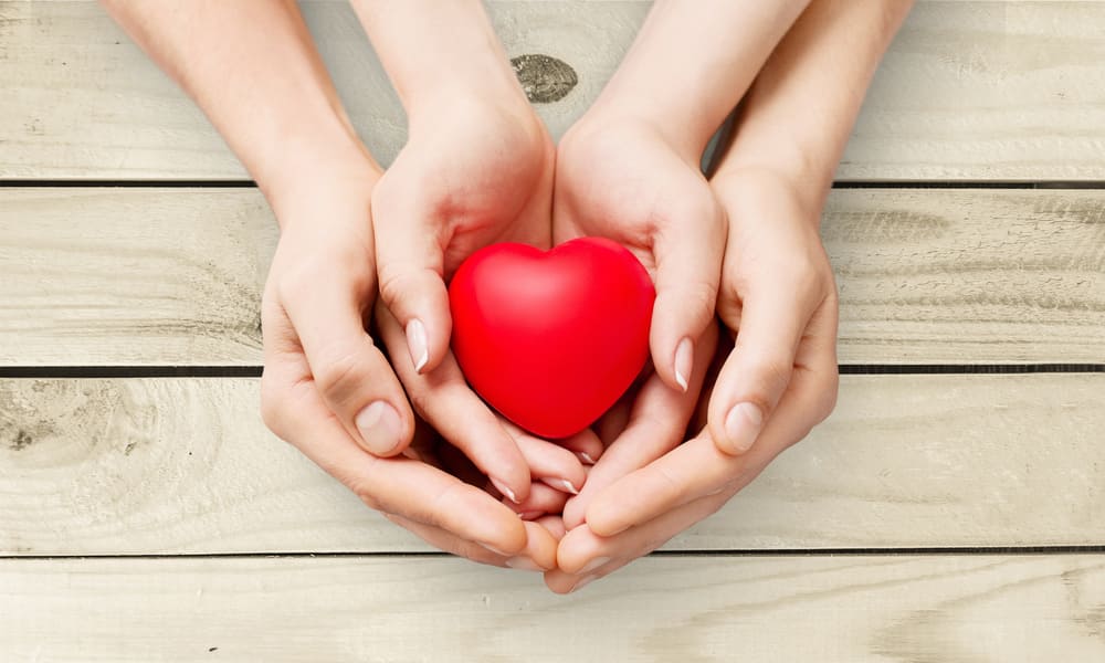 Four hands surrounding a small red heart, symbolizing care and support, placed on a wooden surface—an emblem of various ways to give.