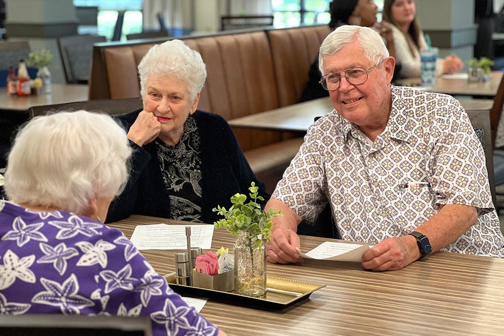 Three elderly people are sitting at a table in a dining area, engaging in conversation. Two women are seated across from an older man who is smiling and holding a paper.