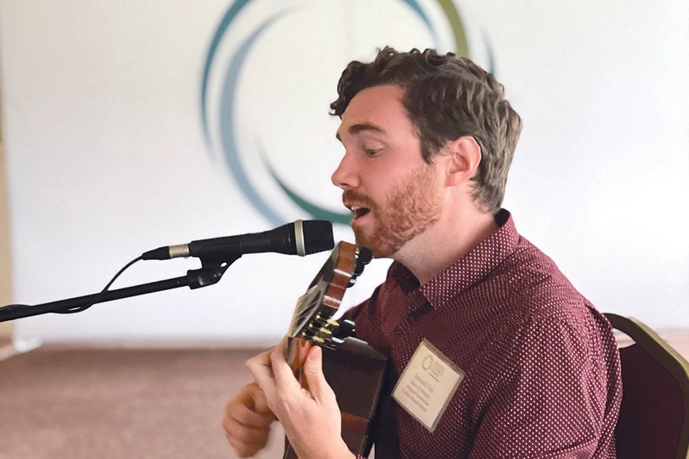 A man in a red shirt sings into a microphone while playing an acoustic guitar in a senior living community. He has short curly hair and a beard, and is wearing a name badge. The background is blurred with a circular design.