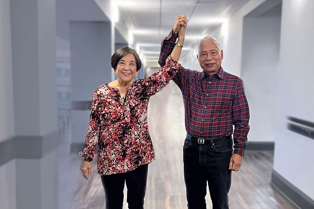 Two elderly adults, one woman and one man, holding hands and raising them in a light hallway at their senior living community. The woman is wearing a floral blouse, and the man is in a plaid shirt.