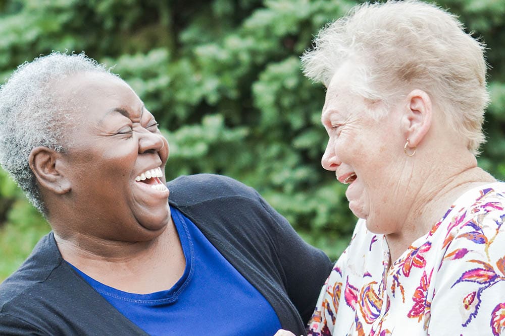 Two older women, enjoying their time in a senior living community, are outdoors laughing together. One wears a blue shirt and black cardigan, while the other sports a patterned blouse. Lush trees provide a serene backdrop to their joyful moment.