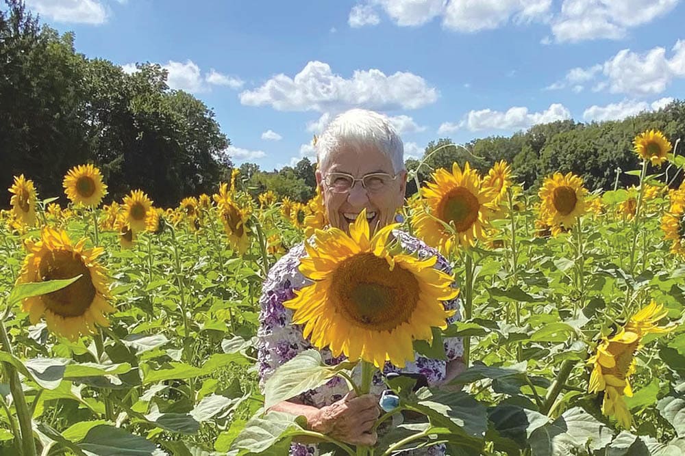 An elderly person stands in a sunflower field, smiling while holding a sunflower. The sky is bright with scattered clouds, and trees line the background. This moment of joy highlights the beauty of senior living and the peaceful environment that surrounds skilled nursing facilities.